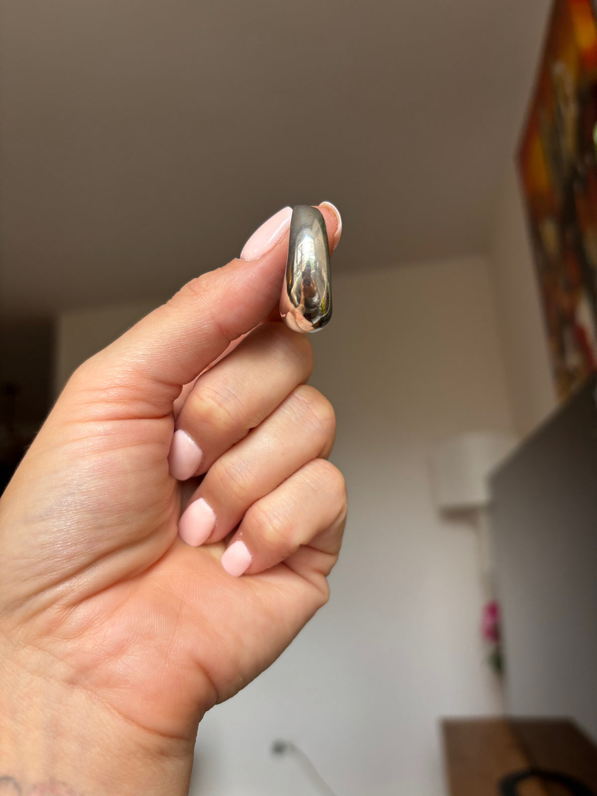 A hand with light pink manicured nails holds a small, polished silver ring against a blurred indoor background.