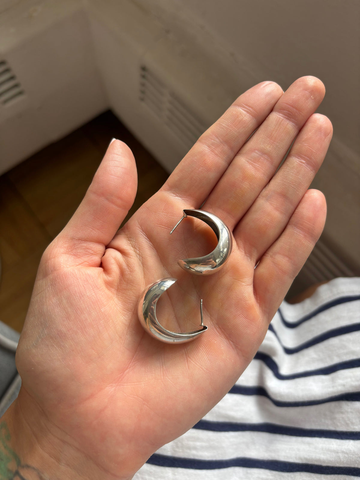 A person's hand holding a pair of polished silver crescent-shaped hoop earrings.