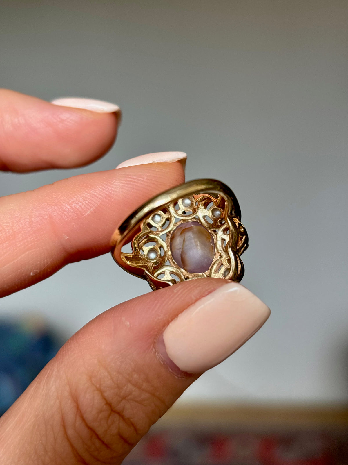 A close-up of a hand holding a gold filigree ring with a central oval stone.