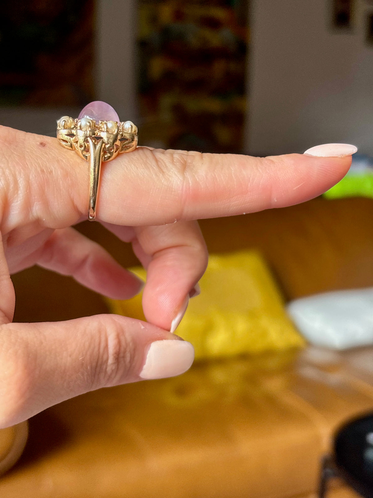 A gold ring with a large oval pink gemstone and smaller side stones, worn on a person's finger in a warm indoor setting.